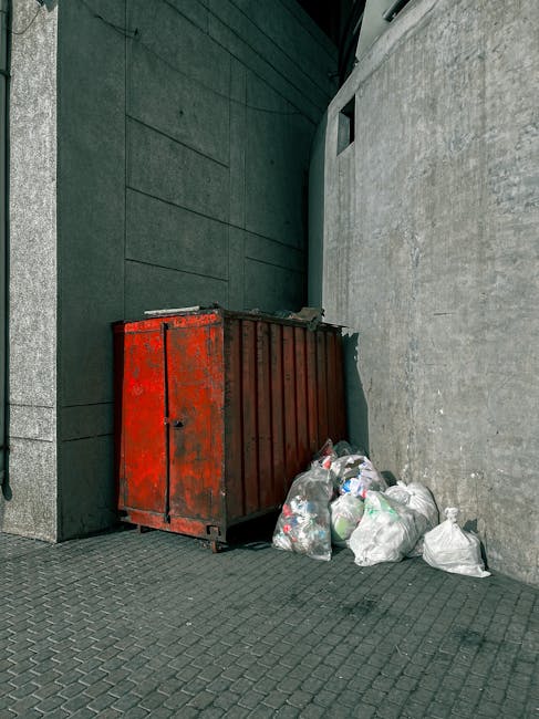 A large outdoor scene showing a collection of stacked cardboard fruit and vegetable boxes labeled 'fresh fruits,' alongside various types of waste containers including three large green wheelie bins w