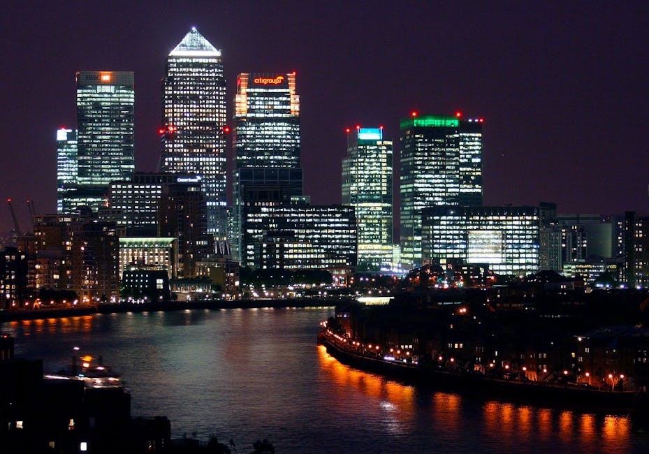 A panoramic view of a city skyline featuring modern high-rise buildings with reflective glass facades, situated behind a row of older, low-rise brick and white-painted structures along the waterfront.