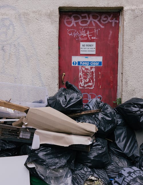Aqueous scene outside a building showing a collection of black rubbish bags piled in front of a weathered brick wall, with some loose cardboard and debris partially visible in the foreground. Behind t